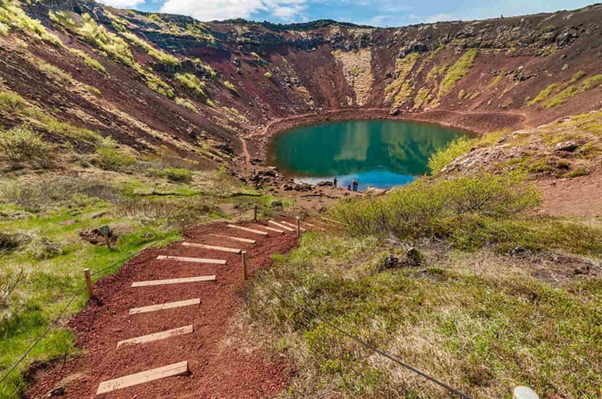 Un sentier bordé de marches mène au cratère de Kerið, dont le lac au centre reflète un vert éclatant, entouré d'une végétation verdoyante et de parois volcaniques rouges.
