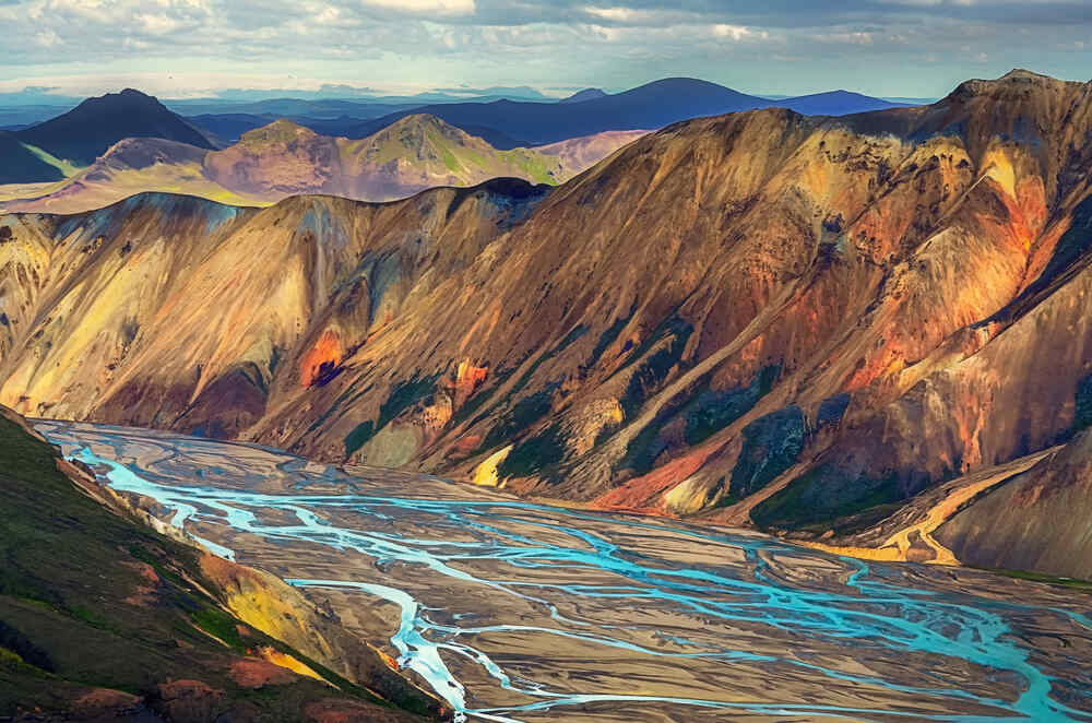 Une vue panoramique des montagnes multicolores de Landmannalaugar, traversées par des rivières sinueuses aux eaux claires, encadrées par des sommets majestueux sous un ciel clair.