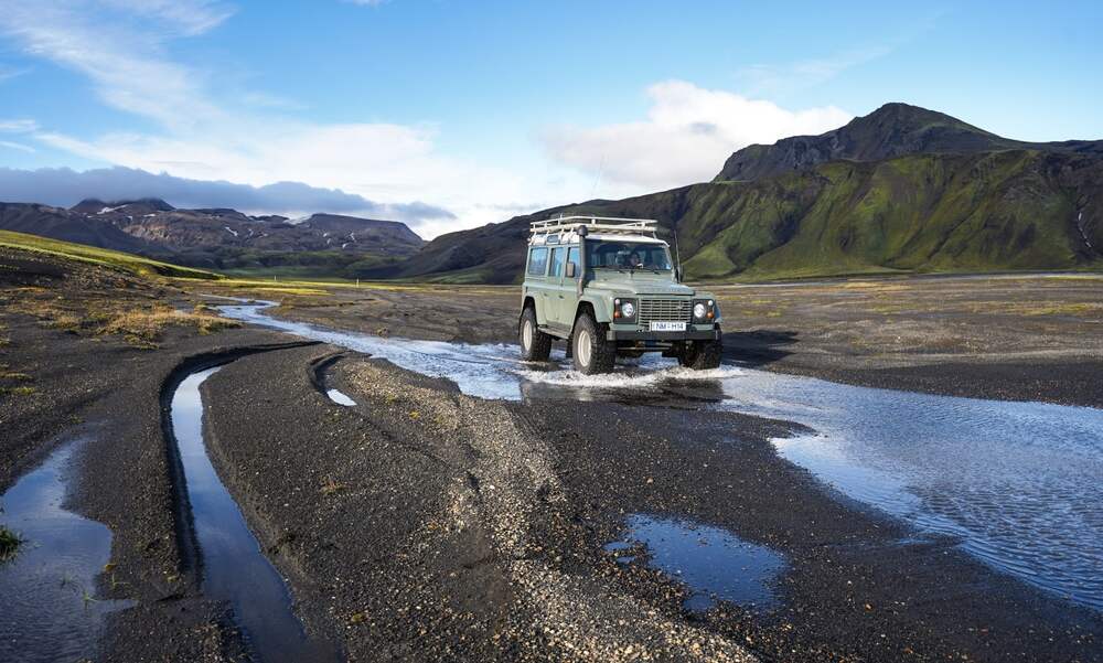 Un véhicule tout-terrain traverse un ruisseau dans les vastes plaines de Landmannalaugar, entouré de montagnes verdoyantes et de paysages volcaniques sous un ciel bleu éclatant.