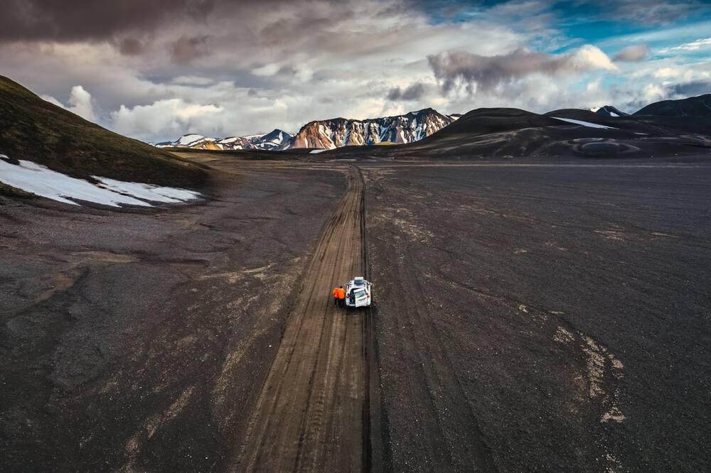 Une voiture tout-terrain traversant une rivière dans la vallée de Landmannalaugar, entourée de montagnes impressionnantes et de vastes étendues de nature sauvage sous un ciel ensoleillé.