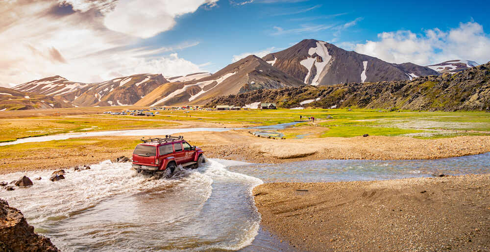 Un véhicule tout-terrain rouge traverse un cours d'eau peu profond dans une vallée entourée de montagnes aux sommets enneigés à Landmannalaugar, sous un ciel dégagé et ensoleillé.