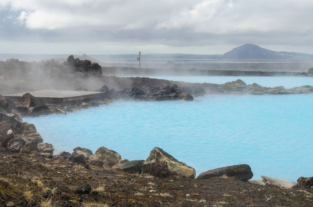 Un bassin d'eau thermale bleu laiteux, entouré de roches volcaniques sombres. Le ciel est couvert de nuages gris, renforçant le contraste avec la teinte éclatante de l'eau.