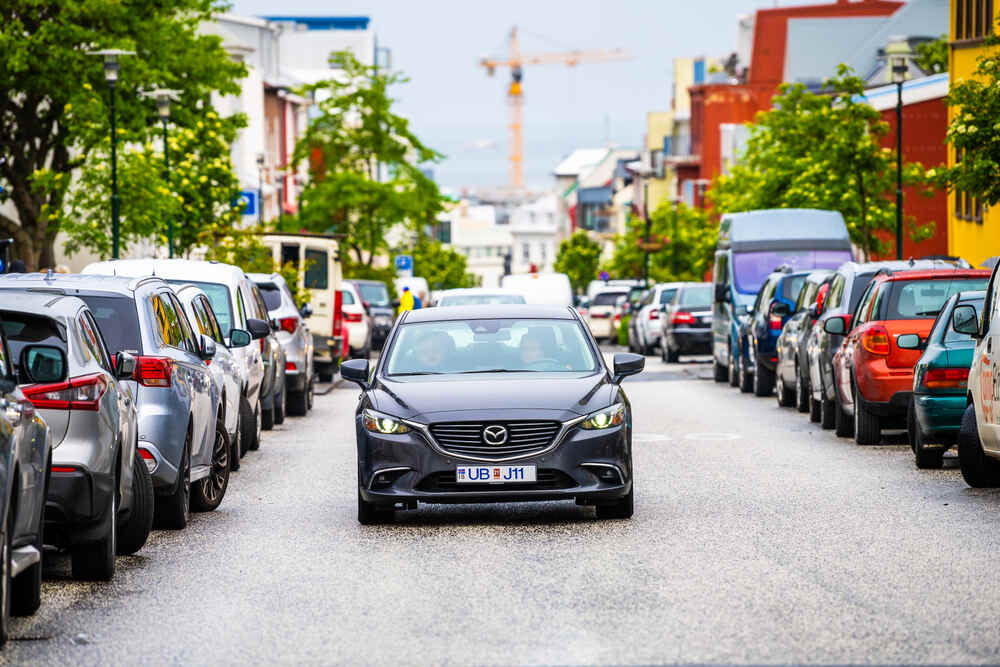 Se garer à Reykjavik Une Mazda noire roulant au centre d’une rue urbaine bordée de voitures garées. Des arbres verts et des bâtiments colorés animent la scène.