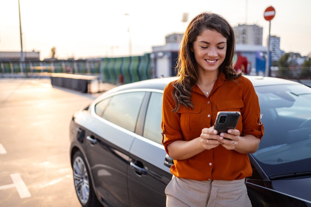 Applications de stationnement à Reykjavik Une femme souriante portant une chemise orange utilise son téléphone portable près d’une voiture moderne, dans un parking ensoleillé.