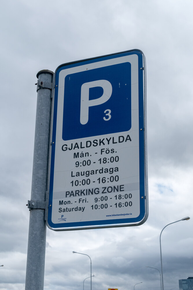 Coût du stationnement à Reykjavik Un panneau de stationnement bleu et blanc à Reykjavik indiquant les horaires et les règles de stationnement, en islandais et en anglais, sous un ciel nuageux.