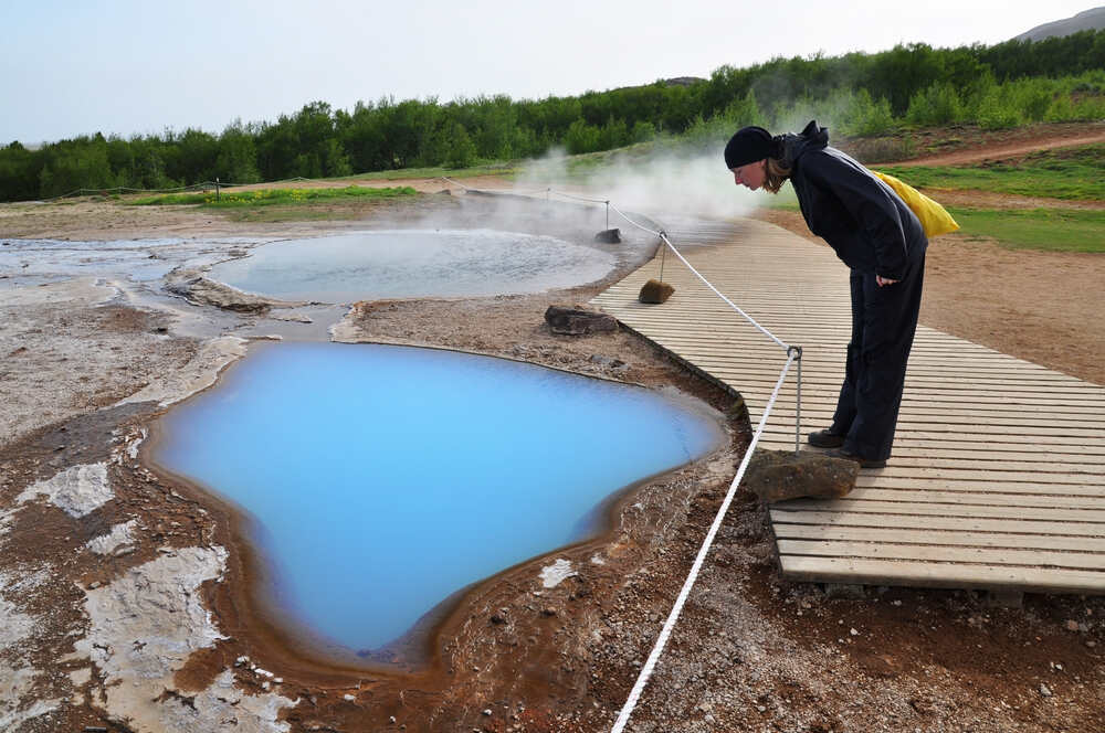 Randonnée et baignade dans les sources thermales d’Islande: la vallée de Reykjadalur Une personne penchée sur une passerelle en bois observe une source thermale bleue lumineuse entourée de vapeur, située dans un paysage naturel. Une corde blanche délimite l’accès pour la sécurité.