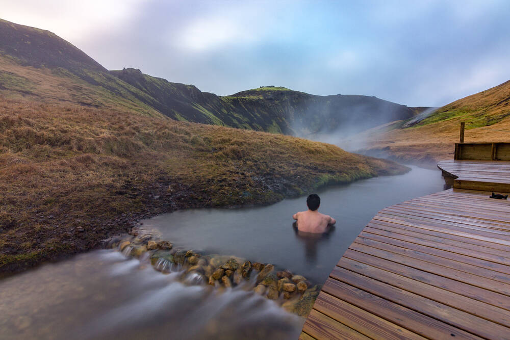 La rivière chaude de Reykjadalur en Islande Un homme assis dans une rivière chaude entourée de collines verdoyantes, avec de la vapeur s’élevant dans l’air. Une passerelle en bois mène jusqu’à l’eau, offrant un accès aux visiteurs.
