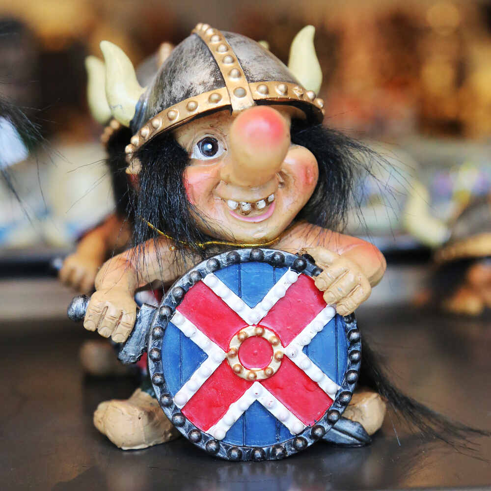 Une figurine humoristique représentant un troll islandais avec un casque viking et un bouclier décoré aux couleurs d’un drapeau croisé rouge et bleu. Le troll arbore un sourire espiègle et un nez proéminent.