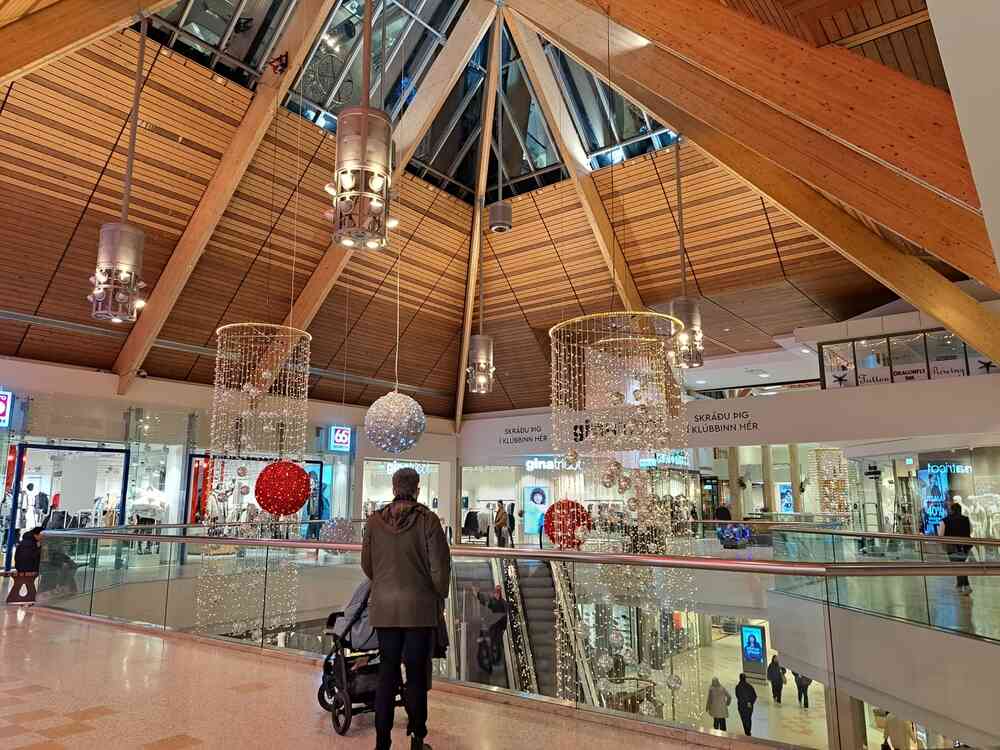 Intérieur d'un centre commercial moderne en Islande, illuminé par de grandes décorations suspendues en forme de sphères lumineuses et entouré de boutiques. Un plafond en bois incliné ajoute une atmosphère chaleureuse et accueillante.