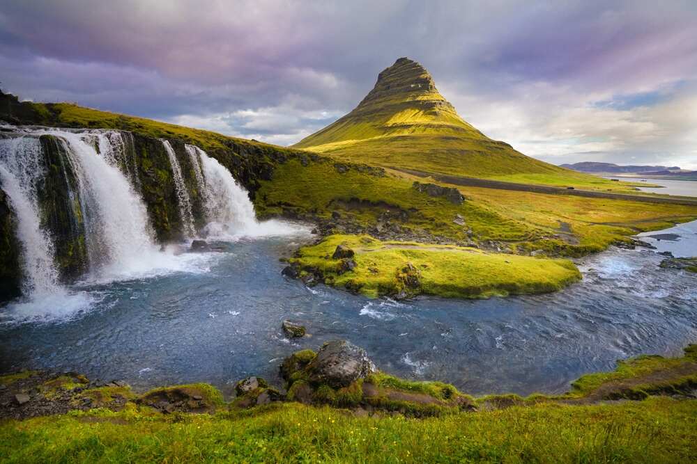 Une majestueuse cascade coule devant une montagne conique emblématique appelée Kirkjufell. Le paysage est verdoyant avec un ciel légèrement nuageux, ce qui ajoute une atmosphère mystique à la scène.