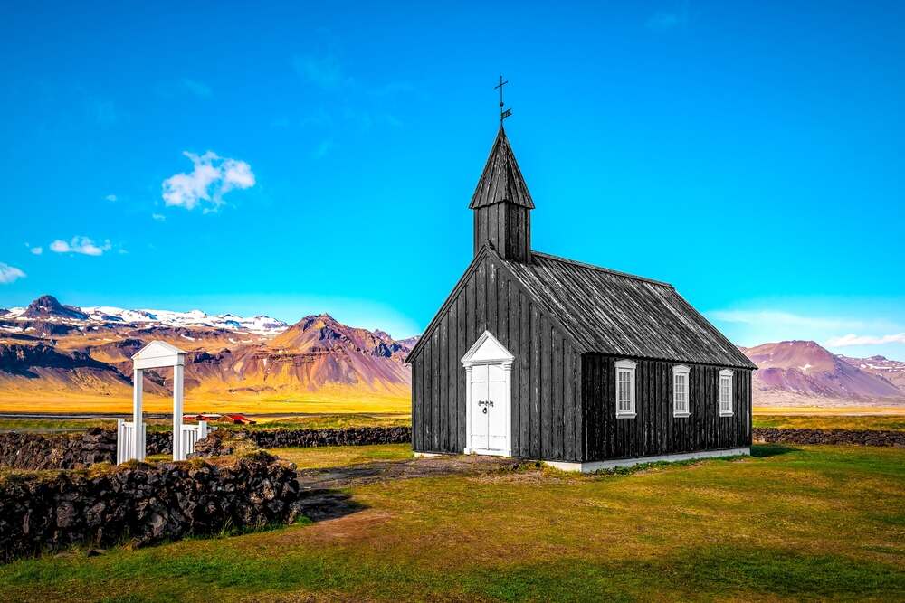Une petite église en bois noire se tient fièrement au milieu d'un paysage islandais spectaculaire, entourée de montagnes et d'un ciel bleu éclatant. L'église contraste avec l'herbe verte et les collines environnantes.