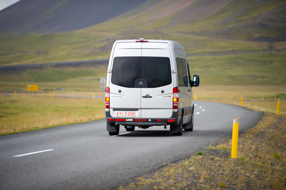 Minifourgonnette Une camionnette blanche roulant sur une route asphaltée dans une vallée islandaise, entourée de collines vertes sous un ciel couvert.