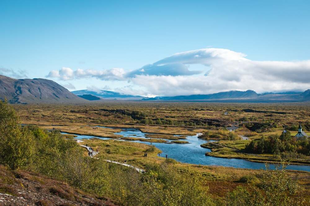 Le parc national de Thingvellir Un paysage à couper le souffle dans le parc national de Thingvellir, avec une rivière traversant une vaste plaine entourée de montagnes au loin sous un ciel bleu clair.