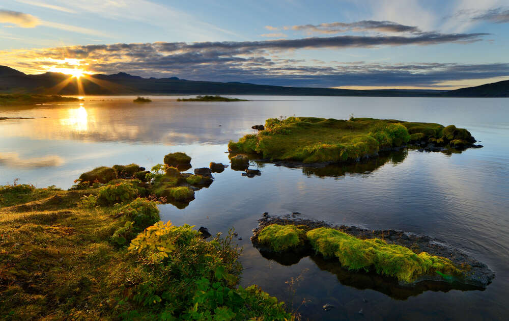 Pêche au lac Thingvallavatn Coucher de soleil sur le lac Thingvallavatn, avec le soleil se reflétant sur l'eau calme, les petites îles couvertes de mousse et un ciel partiellement nuageux.