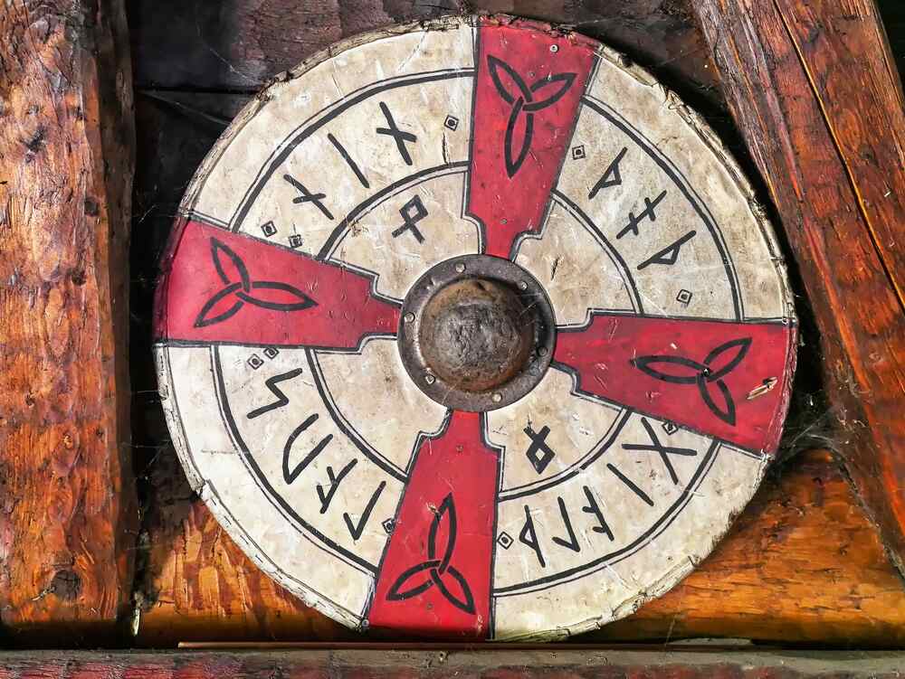 Aegishjalmur Un grand bouclier circulaire en bois dont les parties sont peintes en rouge et en blanc, décoré de runes et de symboles nordiques. Au centre se trouve un boîtier métallique rond.