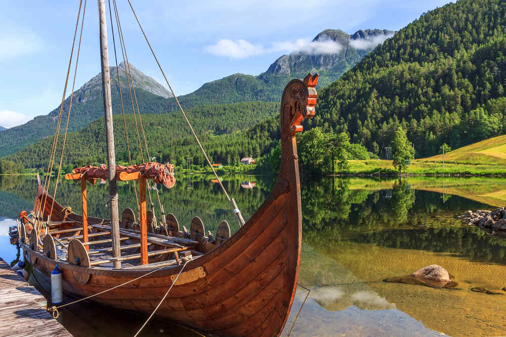 Un bateau viking en bois avec une tête de dragon sculptée dans la proue, amarré sur un lac tranquille entouré de montagnes verdoyantes et d'un ciel clair.