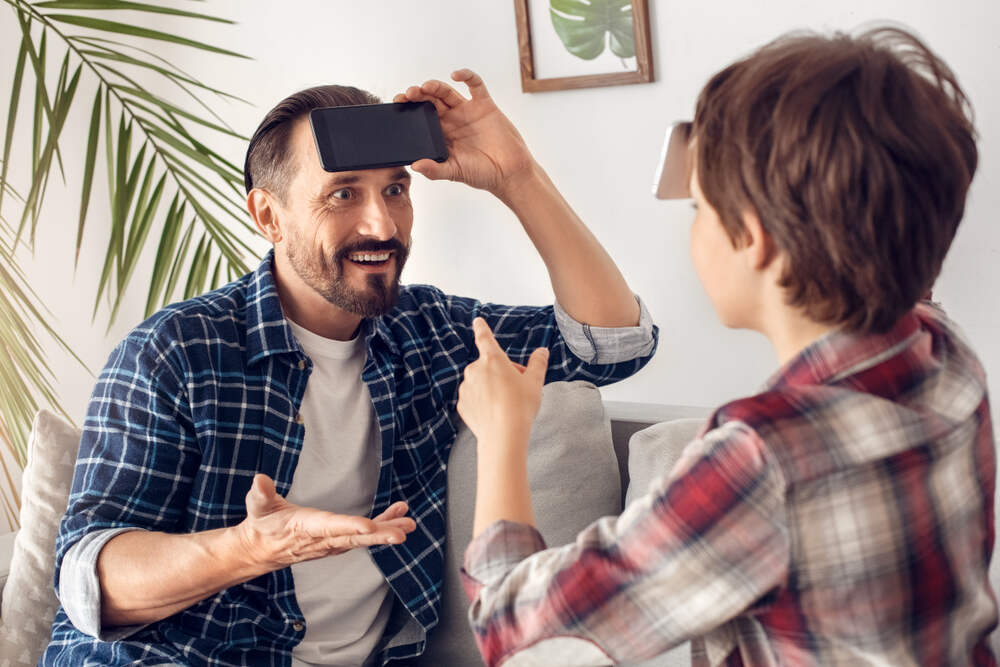 Un homme portant une chemise à carreaux joue à un jeu de devinettes avec un enfant. L'homme tient un smartphone sur son front, tandis que l'enfant lui donne des indices en pointant du doigt avec enthousiasme.