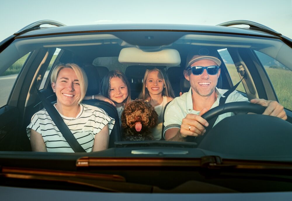 Une famille souriante avec deux enfants et un chien à bord d'une voiture, tous attachés et prêts pour une aventure.
