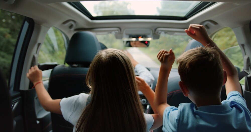 Deux enfants s'amusent sur les sièges arrière d'une voiture en levant les bras, avec une vue sur la route à travers le pare-brise.