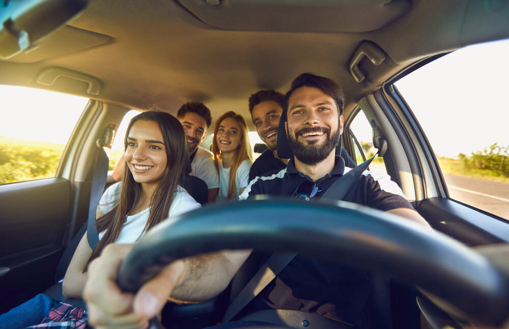 Un groupe de cinq amis souriants prend un selfie depuis l'intérieur d'une voiture en mouvement, le conducteur tenant le volant d'une main et le téléphone de l'autre.