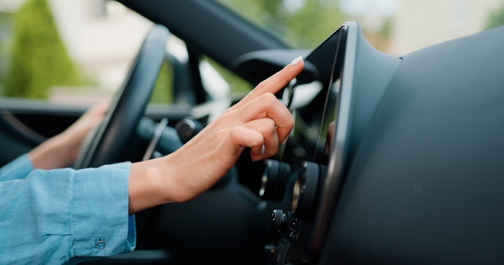 Une main féminine touche un écran de tableau de bord dans une voiture, indiquant une interaction avec un système de navigation ou multimédia.