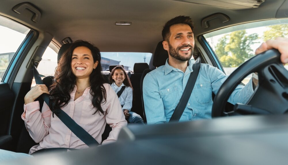 Un couple souriant conduisant avec leur enfant à l'arrière, profitant d'un trajet en voiture.
