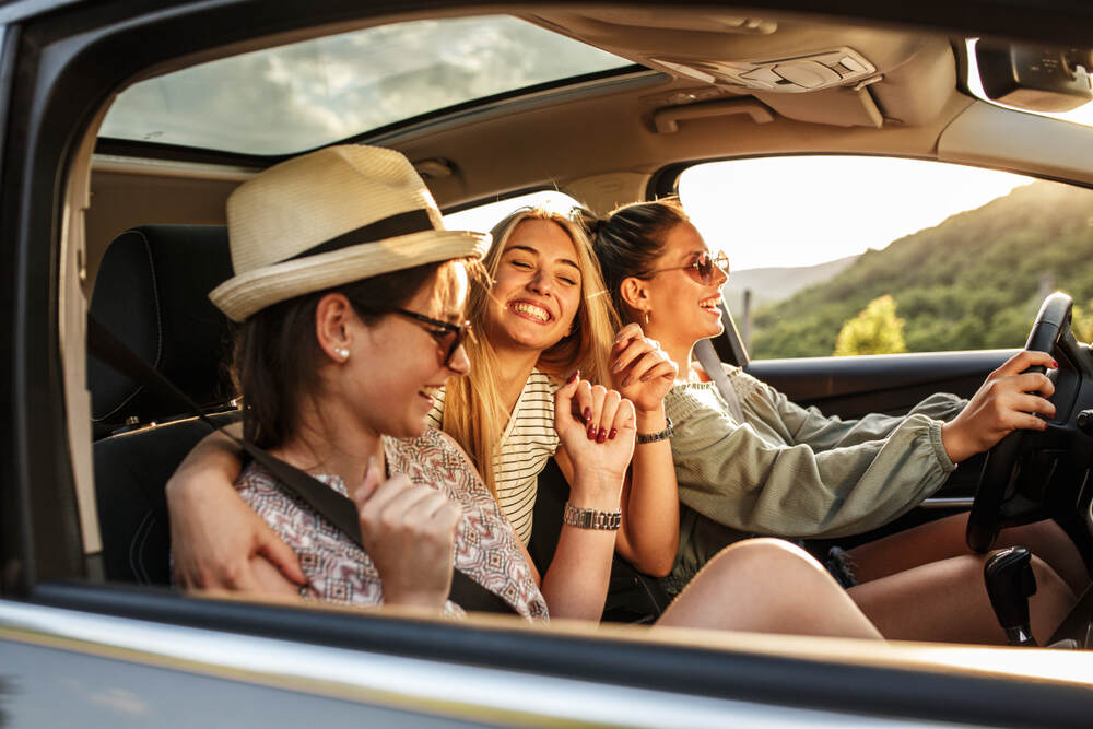 Trois jeunes femmes souriantes dans une voiture, la conductrice portant des lunettes de soleil et un chapeau, pendant une balade sous un ciel ensoleillé.