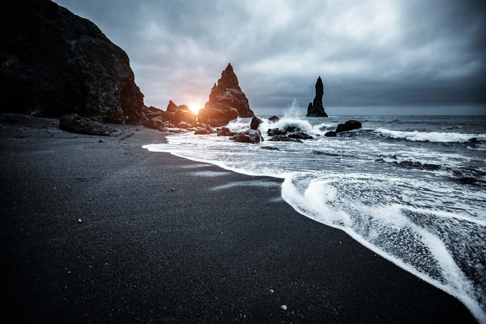 Vue au crépuscule d'une plage de sable noir avec des vagues blanches qui avancent doucement vers le rivage. Les formations rocheuses de Reynisdrangar se dressent majestueusement à l'horizon, tandis qu'un rayon de soleil perce les nuages sombres.