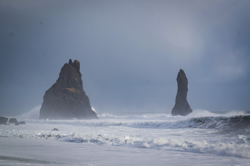 Deux piliers de roche imposants surgissent des vagues déchaînées sous un ciel gris orageux. L'eau s'écrase avec puissance contre la plage de sable noir, créant une atmosphère dramatique et sauvage.