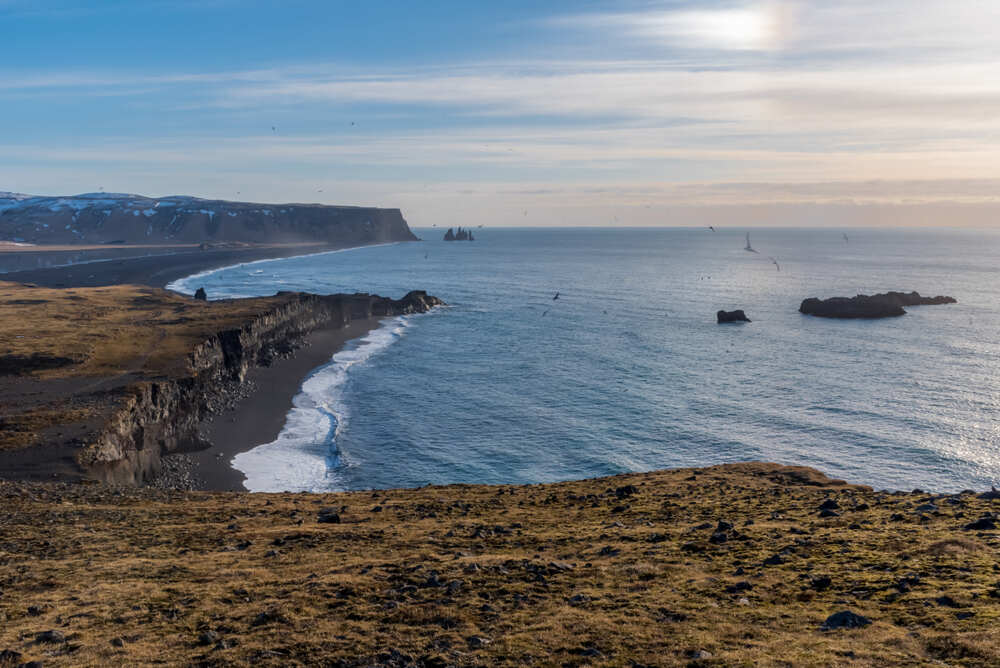 Une vue panoramique d'une falaise islandaise surplombant l'océan Atlantique, avec des vagues se brisant sur la côte et les célèbres pics rocheux Reynisdrangar visibles au loin. Le ciel est bleu clair avec une touche de lumière douce.