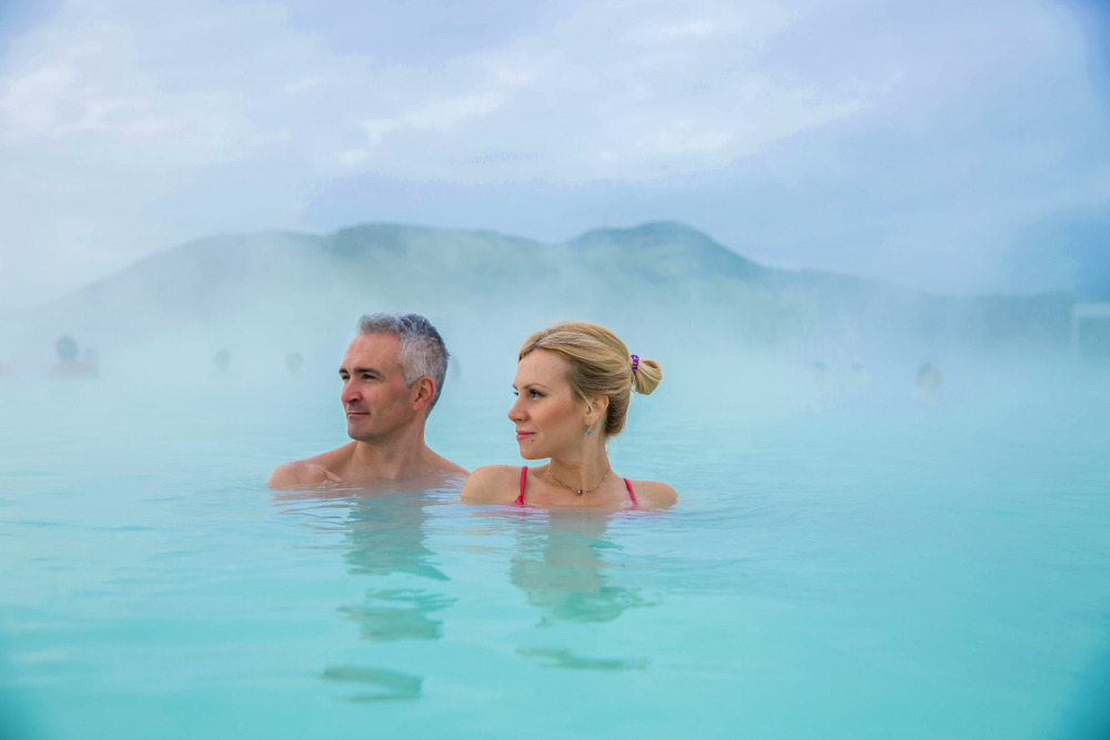 Un couple se détend dans les eaux bleues et fumantes du Blue Lagoon en Islande, entouré de brume qui crée une atmosphère relaxante et mystérieuse.