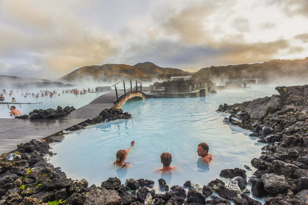 Vue panoramique d'une famille se baignant dans le Blue Lagoon avec des rochers noirs de lave en premier plan et un pont en bois enjambant l'eau fumante sous un ciel nuageux.