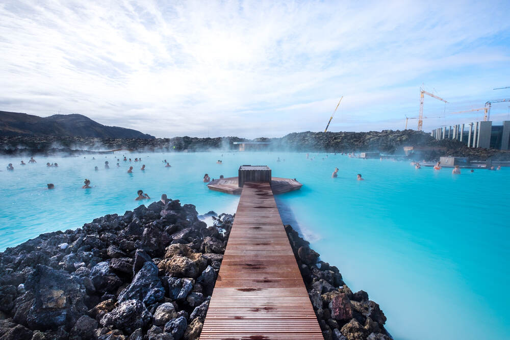 Un ponton en bois s'étend vers les eaux bleu turquoise du Blue Lagoon, entouré de rochers volcaniques noirs, avec plusieurs baigneurs profitant du bain géothermal.