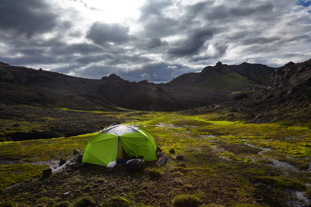 Une tente verte est installée au milieu d'un paysage volcanique. Le sol est tapissé de mousse verte, et les montagnes sombres encerclent l’endroit. Le ciel est couvert de nuages gris, laissant apparaître un éclat de lumière à l'horizon.