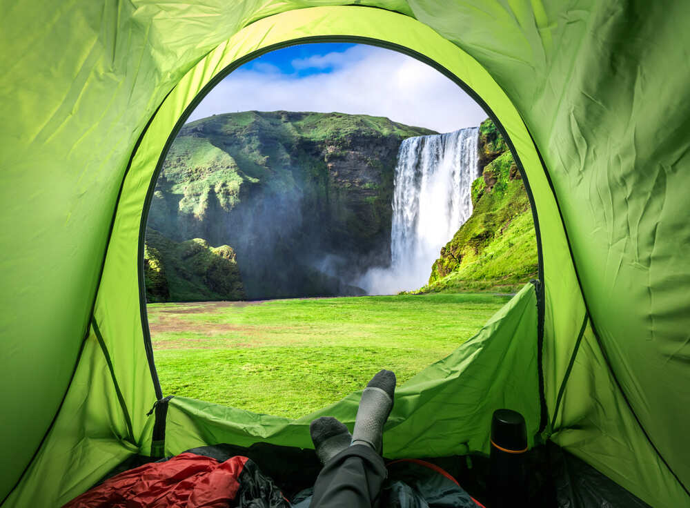Vue de l'intérieur d'une tente verte donnant sur une cascade impressionnante entourée de collines verdoyantes. Les pieds d'une personne reposent sur un sac de couchage rouge, illustrant une pause contemplative face au paysage naturel.