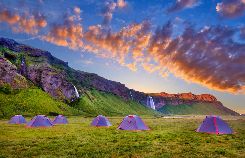 Plusieurs tentes rouges sont alignées sur une vaste plaine herbeuse, entourée de falaises et de cascades. Le ciel est éclatant avec des nuances de bleu et d'orange, annonçant un lever ou un coucher de soleil.