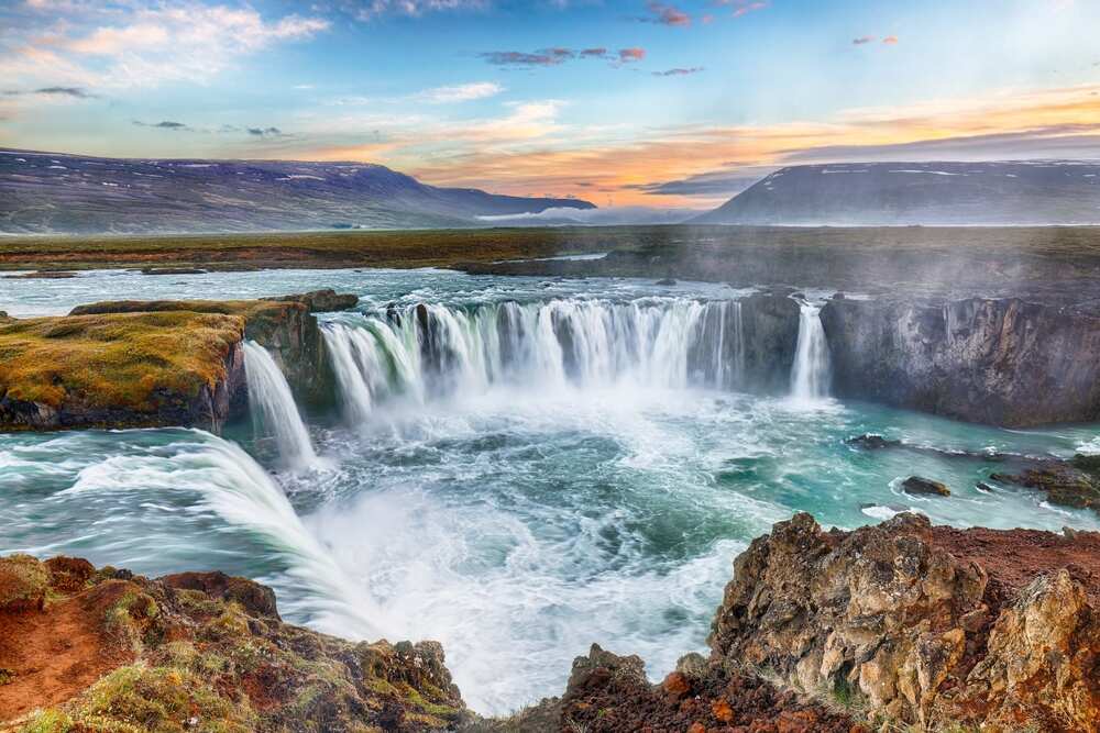 La cascade de Godafoss au coucher du soleil, avec ses puissants torrents d'eau se déversant dans un bassin entouré de paysages verdoyants.