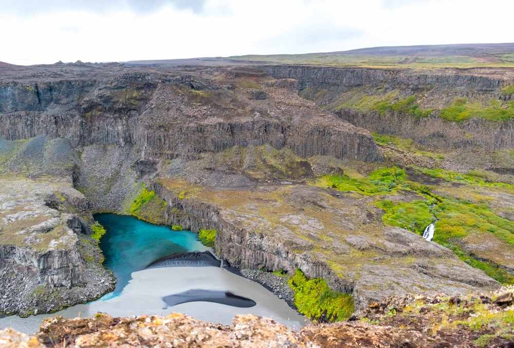 Un paysage spectaculaire de canyon en Islande, avec des falaises de basalte sombre bordant un cours d'eau turquoise qui serpente à travers la roche. 