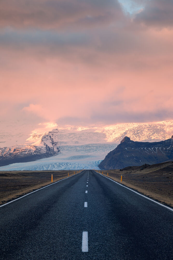 Route islandaise menant aux montagnes glacées, sous un ciel rose et bleu lors du coucher de soleil.