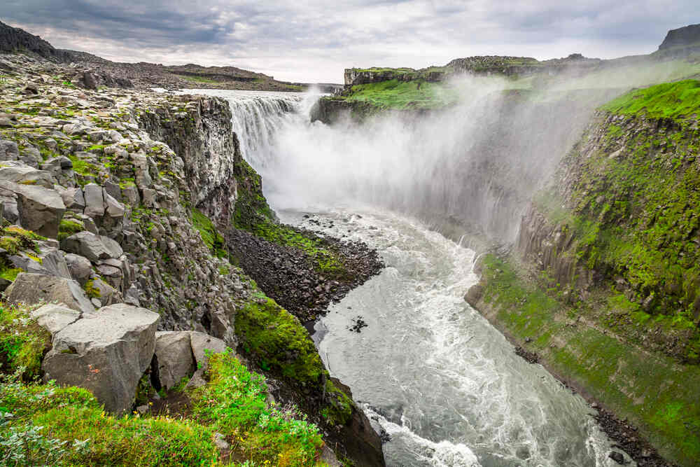 Une imposante cascade appelée Dettifoss, entourée de rochers et de verdure avec une brume épaisse qui s'élève à cause de la puissance de l'eau.