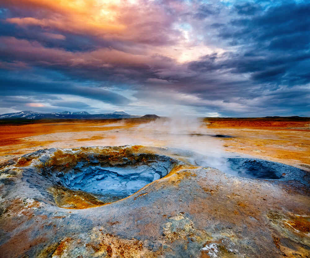 Sol volcanique fumant aux couleurs rougeâtres et grises sous un ciel dramatique aux nuances orange et bleu.