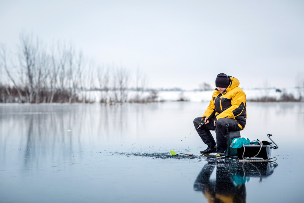 Un pêcheur vêtu d'une veste jaune et noire est assis sur un lac gelé, tenant une canne à pêche près d'un trou creusé dans la glace. L'environnement est enneigé avec des arbres dénudés en arrière-plan, évoquant le calme de l'hiver.