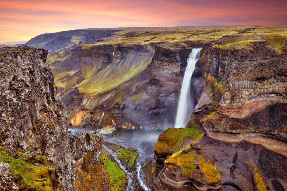 Coucher de soleil sur Haifoss - La majesté de l'une des plus grandes cascades d'Islande. Vue panoramique de la cascade Haifoss sous un ciel aux teintes orangées, avec ses chutes impressionnantes et ses rochers moussus.