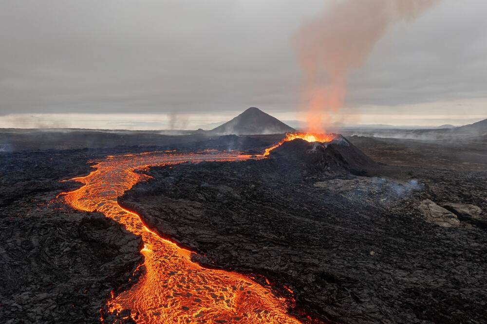 Coulée de lave lors d'une éruption volcanique en Islande. La lave chaude s'écoulant à travers une étendue de roches volcaniques sombres, avec un volcan fumant à l'arrière-plan.