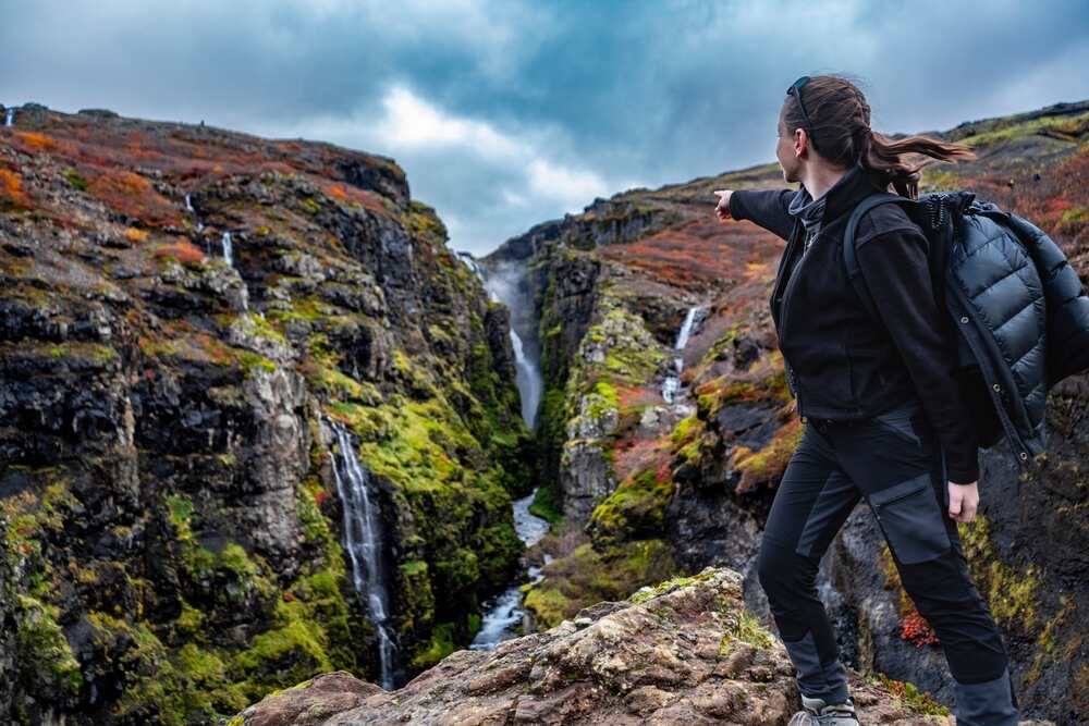 Une randonneuse surplombant un canyon avec plusieurs cascades entourées de végétation automnale aux couleurs vives, pointant vers la chute d'eau principale sous un ciel nuageux.
