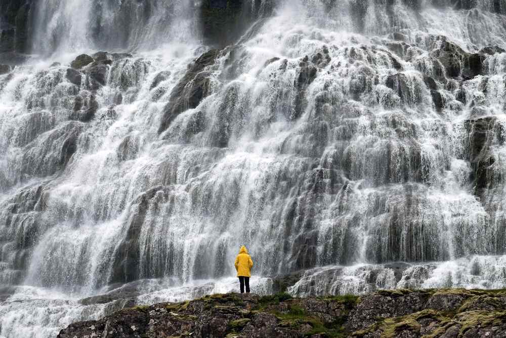 Une personne en manteau jaune se tenant devant la grande cascade de Dynjandi, où l'eau coule en plusieurs niveaux sur une large falaise rocheuse.