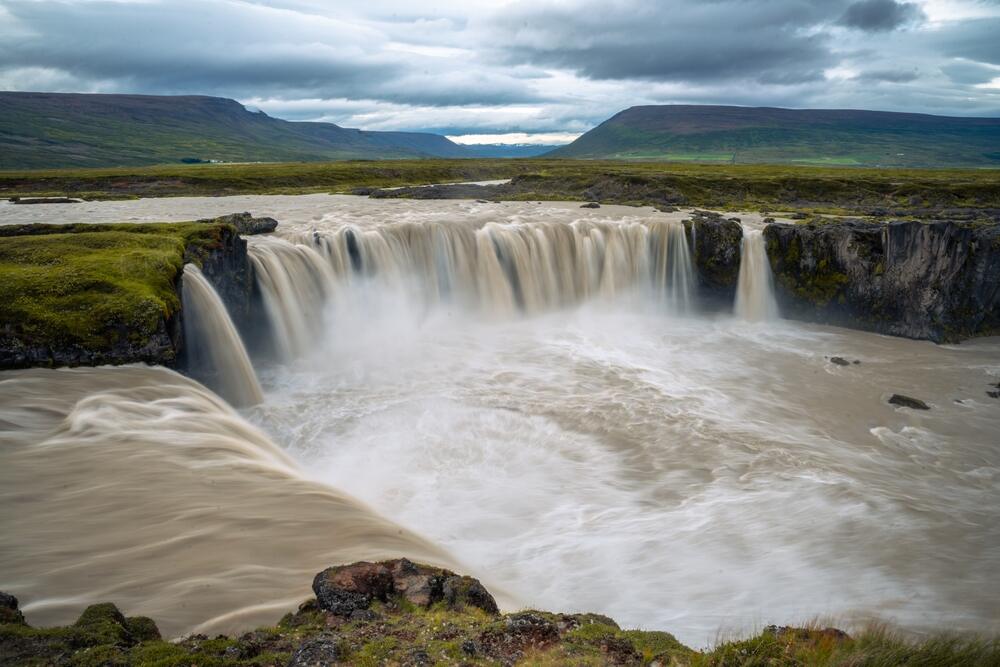 Vue majestueuse de la cascade de Godafoss en Islande avec des eaux tumultueuses entourées de paysages verdoyants et de montagnes en arrière-plan.