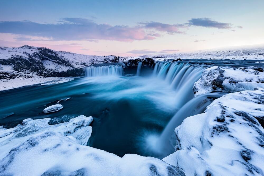 La cascade de Godafoss en hiver, entourée de neige immaculée et éclairée par une lumière douce du coucher de soleil. Les eaux bleues contrastent avec la blancheur éclatante de la glace.