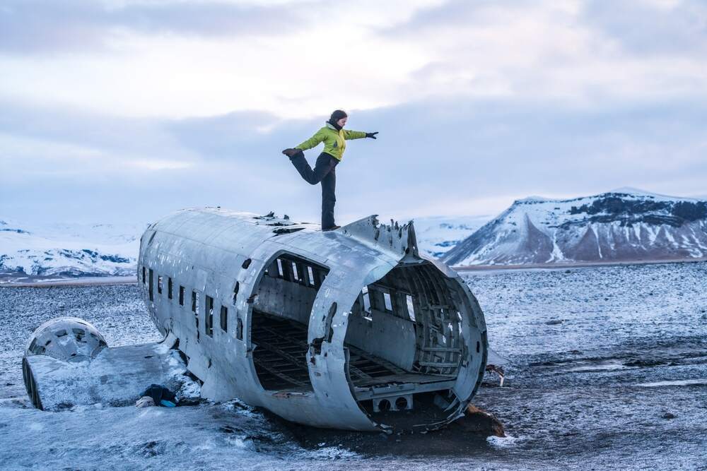 Exploration de l'épave d'avion de Sólheimasandur en Islande Personne debout en équilibre sur l'épave d'avion DC-3 sur la plage de Sólheimasandur avec un décor enneigé à l'arrière-plan.