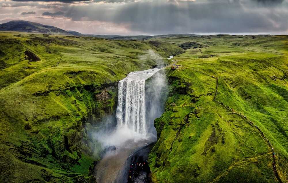 La majestueuse cascade Skógafoss tombant d'une haute falaise verdoyante sous un ciel nuageux.